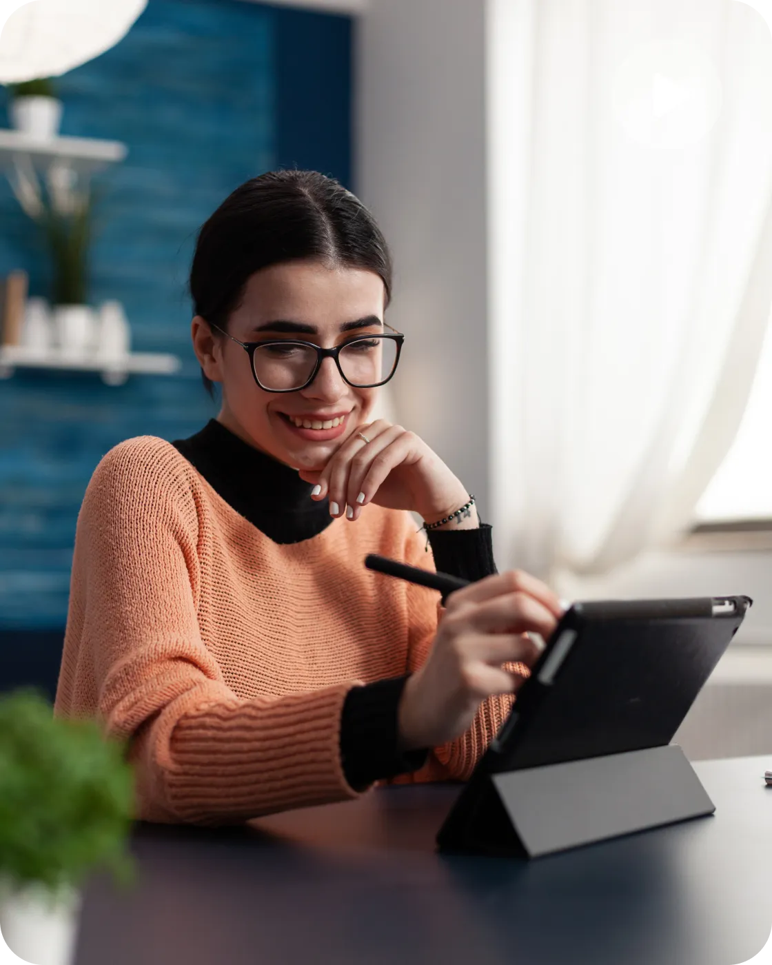 Smiling woman wearing glasses and a peach sweater uses a stylus on a tablet at a desk.