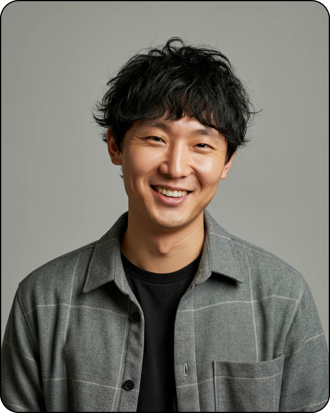 Smiling young man with curly black hair wearing a gray checkered shirt over a black t-shirt on a gray background.