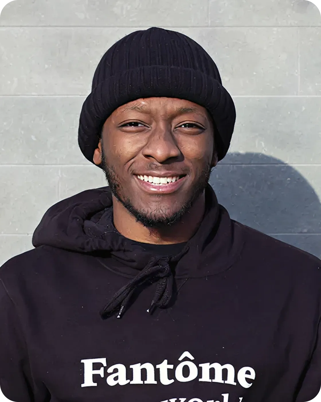 Smiling man wearing a black knit beanie and black hoodie with white text standing against a gray tiled wall.