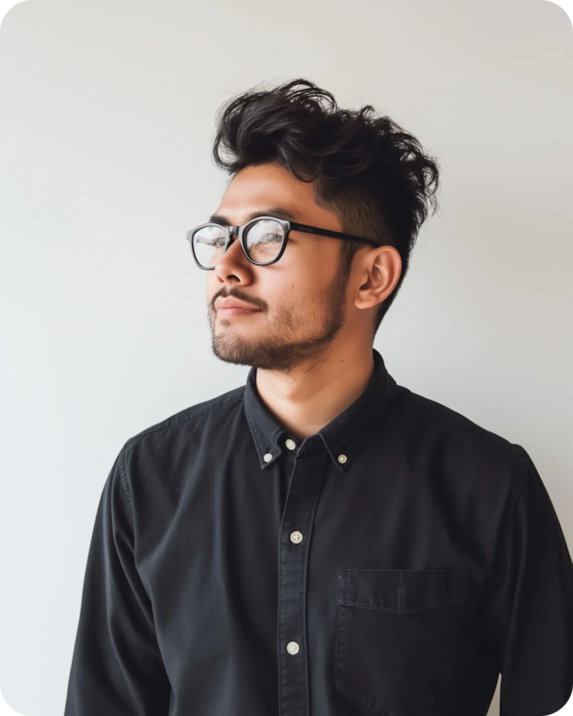 Young man with styled hair and glasses wearing a black button-up shirt looking to the side against a plain background.