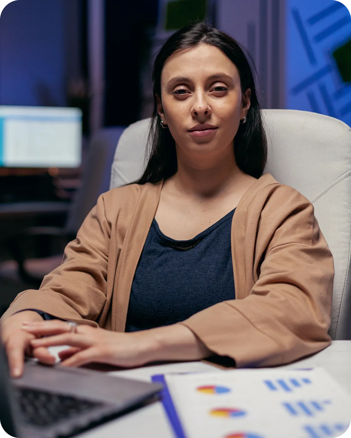 Woman sitting at a desk with a laptop and charts, looking confidently at the camera.