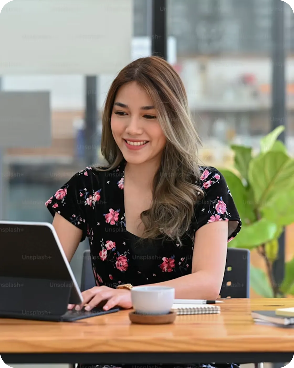 Smiling woman with long hair in a floral dress typing on a tablet at a wooden desk with a cup and notebook.