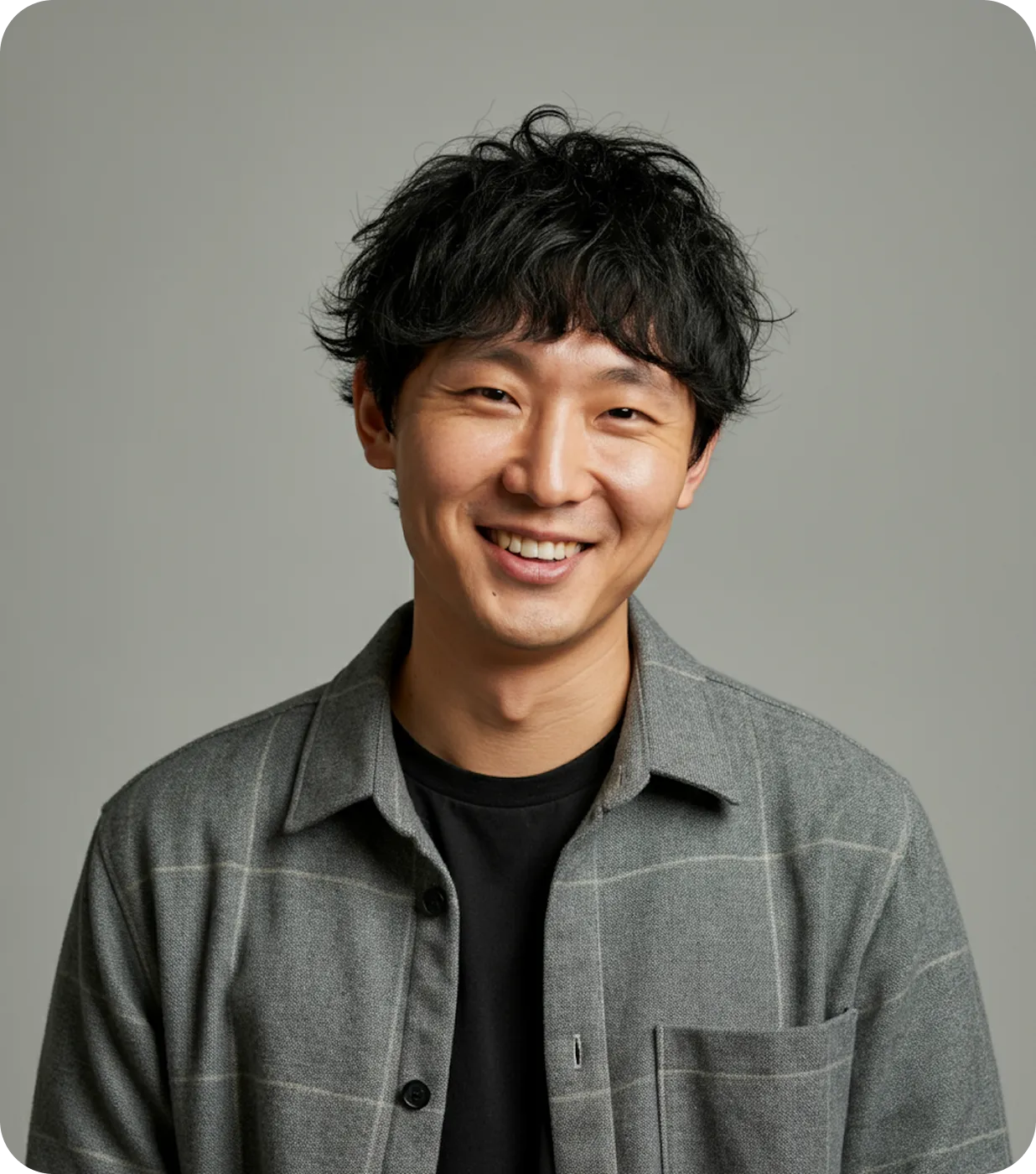 Smiling young man with dark wavy hair wearing a grey checkered shirt over a black t-shirt against a plain grey background.