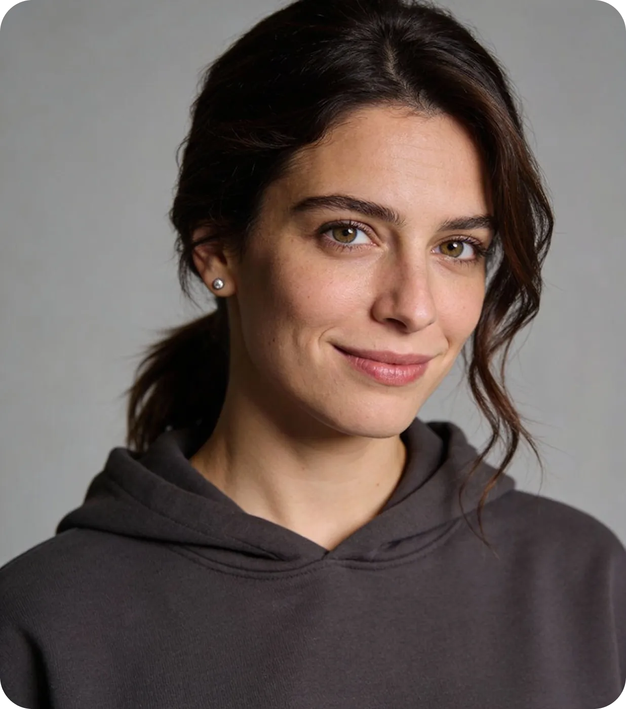 Smiling woman with dark hair in a ponytail wearing a dark hoodie and stud earrings against a plain background.